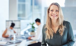 Female leader, manager or CEO with a smile, mission and vision in the office and her team in the background. Leadership, management and success with a woman boss and corporate business people at work